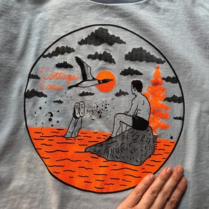 May include: Light blue t-shirt with a circular graphic featuring a loon in flight, an orange sun, and a person sitting on a rock. The words "Cottage Core" are printed above the scene. The shirt is made of a soft material.
