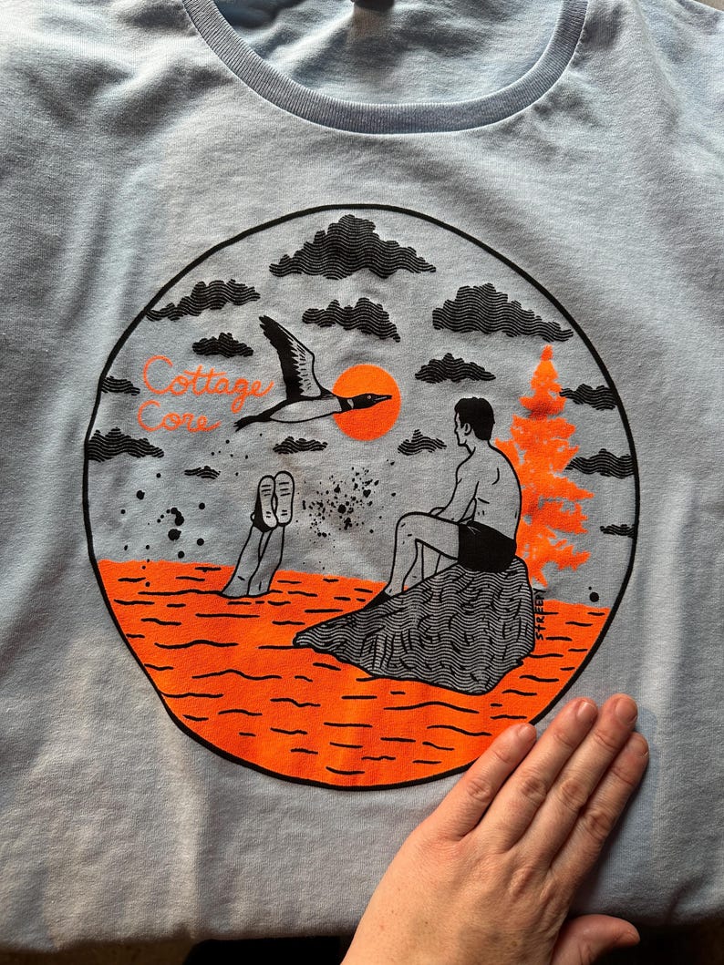 May include: Light blue t-shirt with a circular graphic featuring a loon in flight, an orange sun, and a person sitting on a rock. The words "Cottage Core" are printed above the scene. The shirt is made of a soft material.