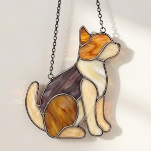 May include: A stained glass suncatcher shaped like a sitting dog, featuring segments of amber, cream, and dark purple glass held together by dark metal solder lines. The piece hangs from a thin metal chain attached to the top of the dog's head.