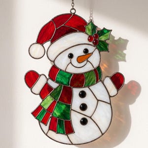 May include: A stained glass snowman ornament featuring a red hat with a white trim, a green and red striped scarf, and three black buttons. The snowman has a bright orange carrot nose, black eyes, and a sprig of green holly with red berries.