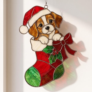 Könnte beinhalten: Ein hängendes Buntglasornament, das einen braun-weißen Welpen mit roter Weihnachtsmannmütze zeigt, der aus einem rot-grünen Weihnachtsstrumpf schaut, verziert mit einer großen roten Schleife und grünen Stechpalmenblättern mit drei kleinen roten Beeren.
