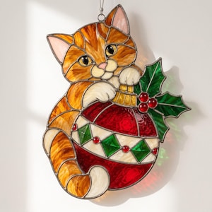 May include: A stained glass suncatcher featuring an orange tabby cat holding a red and white Christmas ornament. The ornament is decorated with green diamond shapes and red berries, accompanied by green holly leaves with a metal frame.