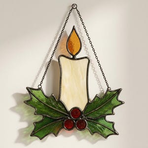 May include: A stained glass hanging ornament featuring a cream-colored candle with an amber flame, surrounded by green holly leaves and three small red berries at the base. The piece is framed in dark metal and suspended by a thin metal chain.