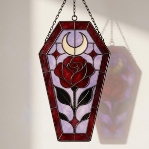 May include: A coffin-shaped stained glass suncatcher featuring a central red rose with green leaves, set against a light purple background. A cream-colored crescent moon sits above the rose, all framed by a deep red border and dark metal cames.