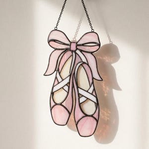 May include: A stained glass suncatcher featuring a pair of ballet slippers in shades of pale pink and cream, tied together with a large pink bow at the top. The piece is outlined in dark metal solder and hangs from a thin black metal chain.