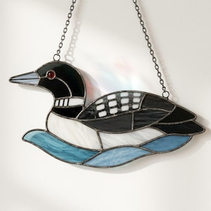 Può includere: Un acchiappasole in vetro colorato a forma di strolaga, composto da segmenti di vetro nero, bianco e blu uniti da giunture in metallo scuro. L'uccello presenta un occhio rosso, una fascia bianca sul collo e un'ala decorata, sospeso tramite una sottile catena metallica.