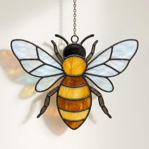 May include: A stained glass bee suncatcher featuring translucent white wings with dark metal leading, a solid black head, and a segmented body with alternating bands of bright yellow and deep amber glass, suspended by a thin metal chain.