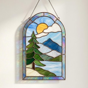 Könnte beinhalten: Ein bogenförmiger Buntglas-Wandbehang mit einer Landschaftsszene, die eine hohe grüne Tanne, ein blaues Gebirge, eine gelbe Sonne und weiße Wolken über einem hellblauen See zeigt, eingefasst von einem dunklen Metallrahmen und einer Aufhängungskette.