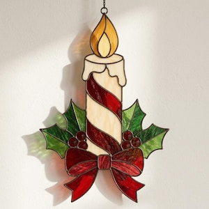May include: A stained glass hanging ornament shaped like a candle with a yellow flame, featuring a cream and red spiral pattern. The base is decorated with green holly leaves, red berries, and a red bow, all outlined in dark metal solder lines.