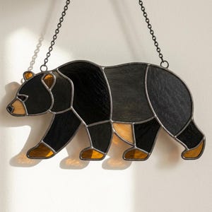 Può includere: Un acchiappasole in vetro colorato a forma di orso che cammina, realizzato con pezzi di vetro nero e ambra uniti da linee di saldatura in metallo scuro. L'orso è sospeso tramite due sottili catene metalliche fissate sulla parte superiore.