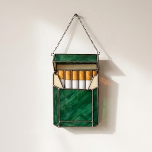 May include: A stained glass wall hanging shaped like an open cigarette pack. The exterior is made of marbled green glass panels with dark metal soldering. Inside, five cylindrical glass pieces with yellow and white tips are visible.