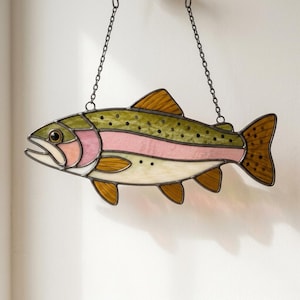 May include: A stained glass suncatcher shaped like a trout fish, featuring olive green, pale pink, and cream-colored glass panels with dark spots. The fish is outlined in dark metal solder and hangs from two thin metal chains attached to its back.
