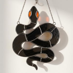 Op de afbeelding: Een glas-in-lood suncatcher in de vorm van een opgerolde slang met zwarte glazen segmenten en feloranje glazen ogen. Het object is geconstrueerd met donkere metalen soldeerlijnen en hangt aan een dunne zilverkleurige metalen ketting.