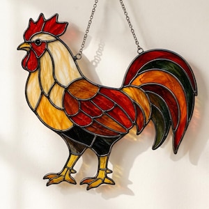 Puede incluir: Un atrapasoles de vidrieras con forma de gallo, que presenta una paleta vibrante de paneles de vidrio rojo, naranja, amarillo y verde delineados con soldadura de metal oscuro. El gallo está suspendido por una fina cadena de metal.