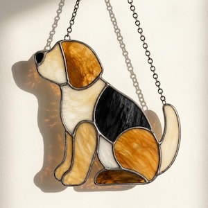 Può includere: Un acchiappasole in vetro colorato a forma di cane seduto, caratterizzato da pezzi di vetro marmorizzato color crema, ambra e nero tenuti insieme da linee di saldatura in metallo scuro. L'oggetto pende da una sottile catena metallica fissata.