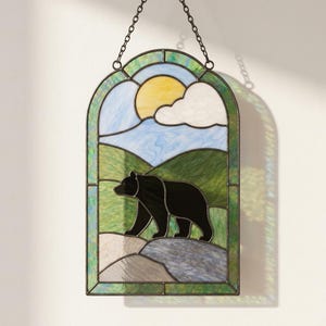 May include: An arched stained glass window hanging featuring a black bear silhouette walking across a rocky landscape. The scene includes green rolling hills, a light blue sky, a yellow sun, and white clouds, all framed by a dark green border.