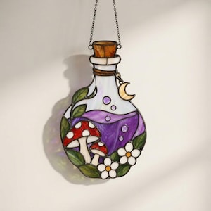 May include: A stained glass suncatcher shaped like a potion bottle with a metal chain. The bottle features a purple liquid interior with bubbles, two red and white mushrooms, green leaves, white flowers, and a small yellow crescent moon charm.