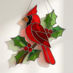 May include: A stained glass suncatcher featuring a bright red cardinal bird with a black face and orange beak, perched on a brown branch surrounded by green holly leaves and clusters of small red berries, suspended by a thin silver metal chain.