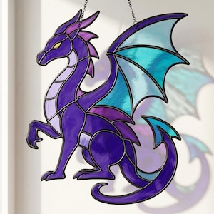 May include: A stained glass suncatcher shaped like a dragon with a deep purple body, light purple accents, and translucent teal wings. The dragon is depicted in a standing profile pose with a long curved tail and yellow eyes, held by a metal chain.