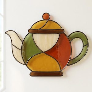 May include: A stained glass teapot wall hanging featuring a multi-colored design with segments of amber, olive green, burnt orange, and cream glass. The pieces are joined by dark metal lead lines, forming a classic teapot shape with a lid and handle.