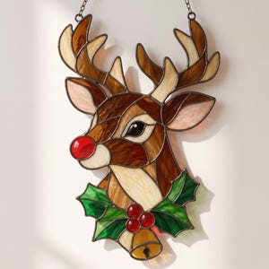 May include: A stained glass suncatcher shaped like a reindeer head with brown, cream, and tan glass pieces. The reindeer has a bright red nose, green holly leaves, red berries, and a small golden bell hanging at the bottom, suspended by a chain.