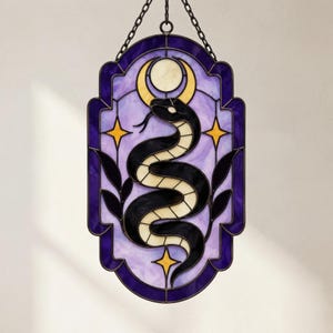 Pode incluir: Um apanhador de sol de vitral retangular apresentando uma cobra preta enrolada sob uma lua crescente amarela e uma lua circular branca. O design inclui estrelas amarelas de quatro pontas e motivos de folhas pretas num fundo roxo claro.