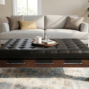 May include: A rectangular black leather tufted ottoman with a dark wood frame featuring horizontal cutouts. A round wooden tray sits on top, holding a white ceramic mug, a clear glass of water, and a small book with a white cover.