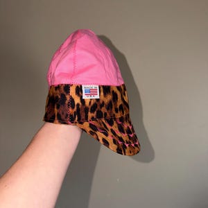 May include: A welding cap featuring a bright pink crown and a leopard print band and brim. The cap includes a small rectangular white label on the front with the text MADE IN U.S.A. and a graphic of the American flag with red, white, and blue colors.