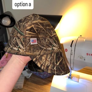 May include: A welding cap featuring a brown, tan, and green camouflage pattern with a dry grass design. The cap has a small rectangular white label sewn on the front that reads "MADE IN U.S.A." with a small American flag graphic above the text.