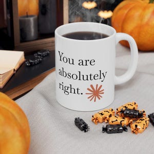 May include: A white ceramic mug with a handle, featuring the black printed text "You are absolutely right." and a small, rust-colored starburst graphic. The mug is filled with a dark liquid and sits on a light-colored fabric surface.