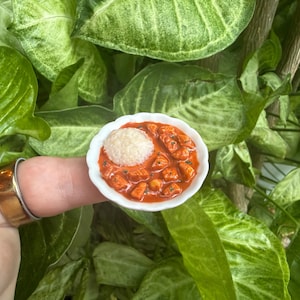May include: A miniature white scalloped bowl containing a serving of orange-red curry with chunks of meat and a single scoop of white rice. The small dish is held on a fingertip, showcasing the tiny scale of the food replica and its details.
