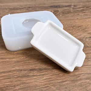 May include: A white rectangular ceramic butter dish lid with small side handles positioned in front of a translucent silicone mold. The lid has a smooth, glossy finish and a recessed center, while the mold features a matching rectangular cavity.