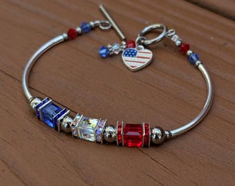 Puede incluir: Un brazalete de plata con cuentas de cristal rojo, blanco y azul y un pequeño dije de bandera estadounidense.