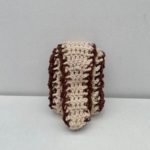 Op de afbeelding: Een ronde, gehaakte honkbal gemaakt van crèmekleurig garen met donkerbruin garen gestikt in een gebogen patroon om het uiterlijk van traditionele honkbalveters na te bootsen. De textuur is zacht en over het hele object zichtbaar.