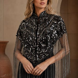 May include: A black bolero jacket featuring a high collar and intricate silver sequin embroidery in a leaf-like pattern. The garment is adorned with long, delicate silver chain fringe that hangs from the hem and sleeves, creating a layered effect.