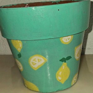 May include: A tapered plant pot painted in a bright turquoise colour, featuring a repeating pattern of yellow lemons and white lemon slices with green leaves. The interior of the pot shows the natural brown terracotta material at the rim.