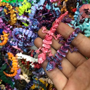Può includere: Una collezione di statuette di lucertole articolate in vari colori, tra cui rosa, blu, viola, giallo, arancione e bianco. Le lucertole presentano un design del corpo segmentato con una texture stampata in 3D visibile e sono in un mucchio.