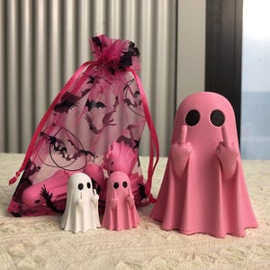 Op de afbeelding: Een grote roze spookfiguur met zwarte ogen en opgestoken middelvingers, vergezeld door twee kleinere spookfiguren in wit en roze, en een roze organza trekkoordzakje met een zwart vleermuispatroon en extra kleine spookfiguren.