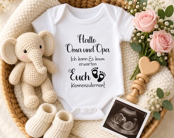 May include: A white baby bodysuit featuring black text that reads Hallo Oma und Opa Ich kann Es kaum erwarten Euch Kennenzulernen with two small footprints. Included are a crocheted elephant, cream booties, a wooden heart rattle, and an ultrasound.