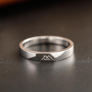 May include: A silver-toned metal ring with a polished finish and a flat, faceted front section. The band features a small, engraved geometric mountain-like symbol centered on the flat surface, creating a clean and simple design.