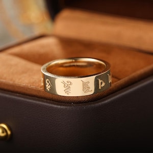 Könnte beinhalten: Ein goldfarbener Bandring mit einer glatten, polierten Oberfläche und eingravierten Schriftzeichen, die den äußeren Rand umschließen. Der Ring hat ein flaches Profil und eine gleichmäßige Breite mit detaillierten, dunkel ausgefüllten Gravuren.