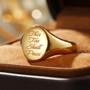 Pode incluir: Um anel de sinete em ouro polido com uma face lisa e arredondada gravada com o texto "This Too Shall Pass" em letra cursiva. O anel tem um acabamento metálico reflexivo e uma banda cónica, apresentando um design limpo e simples.