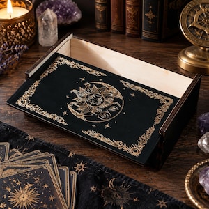 May include: A rectangular wooden box with a black lid featuring a gold-colored sun and moon illustration. The lid is decorated with ornate floral borders and small stars. The interior of the box is light-colored wood, and the exterior sides are dark.