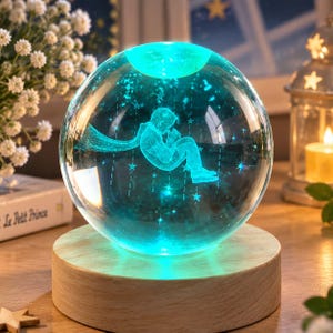 Può includere: Una luce notturna a sfera in vetro trasparente con un'incisione laser 3D interna che raffigura una figura con mantello seduta tra le stelle. La sfera è illuminata da un bagliore turchese e poggia su una base circolare in legno chiaro.
