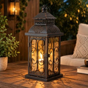 Puede incluir: Un farol rectangular de metal negro con acabado desgastado, que presenta intrincados patrones de volutas en los paneles de vidrio, una puerta con bisagras y un techo decorativo con una pequeña asa, que contiene una vela blanca encendida.