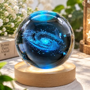 May include: A spherical glass paperweight featuring a deep blue interior with a detailed laser-etched spiral galaxy and small star patterns. The sphere rests on a circular, light-coloured wooden base that provides illumination from below.
