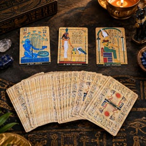 May include: A deck of Egyptian-themed tarot cards with papyrus-style textures. Three cards are displayed face-up, labeled 25 Hapy Renewal, 26 Neith Perseverance, and 27 Satet Fruitfulness, while the remaining cards are fanned out showing hieroglyphic designs.