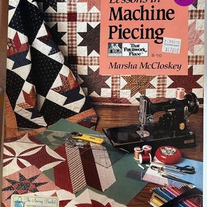May include: A book titled "Lessons in Machine Piecing" by Marsha McCloskey, with a sewing machine, fabric samples, and sewing tools. The cover features a quilt with star patterns. The image includes a price tag and the logo of "That Patchwork Place".
