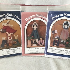 May include: Three vintage craft pattern packets titled "Dream Spinners." The packets feature illustrations of fabric dolls. The packets are labeled "Woodland Choir," "Warm at Heart," and "American Celebration."
