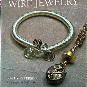 May include: A book titled "Creative Wire Jewelry" with a silver-colored wire bracelet and a pendant with a decorative wire design. The pendant has a gold-colored bead and a dark, speckled stone. The book cover is gray.